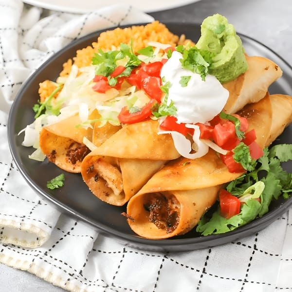 Crispy taquitos with rice, shredded lettuce, diced tomatoes, sour cream, and guacamole on a black plate.
